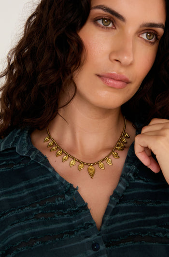 Woman with wavy dark hair wears a blue textured blouse and the Paisley Necklace, a gold-tone accessory inspired by Indian metalwork, looking slightly to the side.
