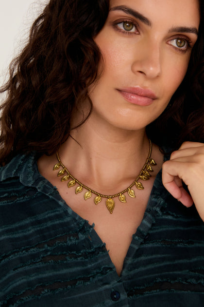 Woman with wavy dark hair wears a blue textured blouse and the Paisley Necklace, a gold-tone accessory inspired by Indian metalwork, looking slightly to the side.