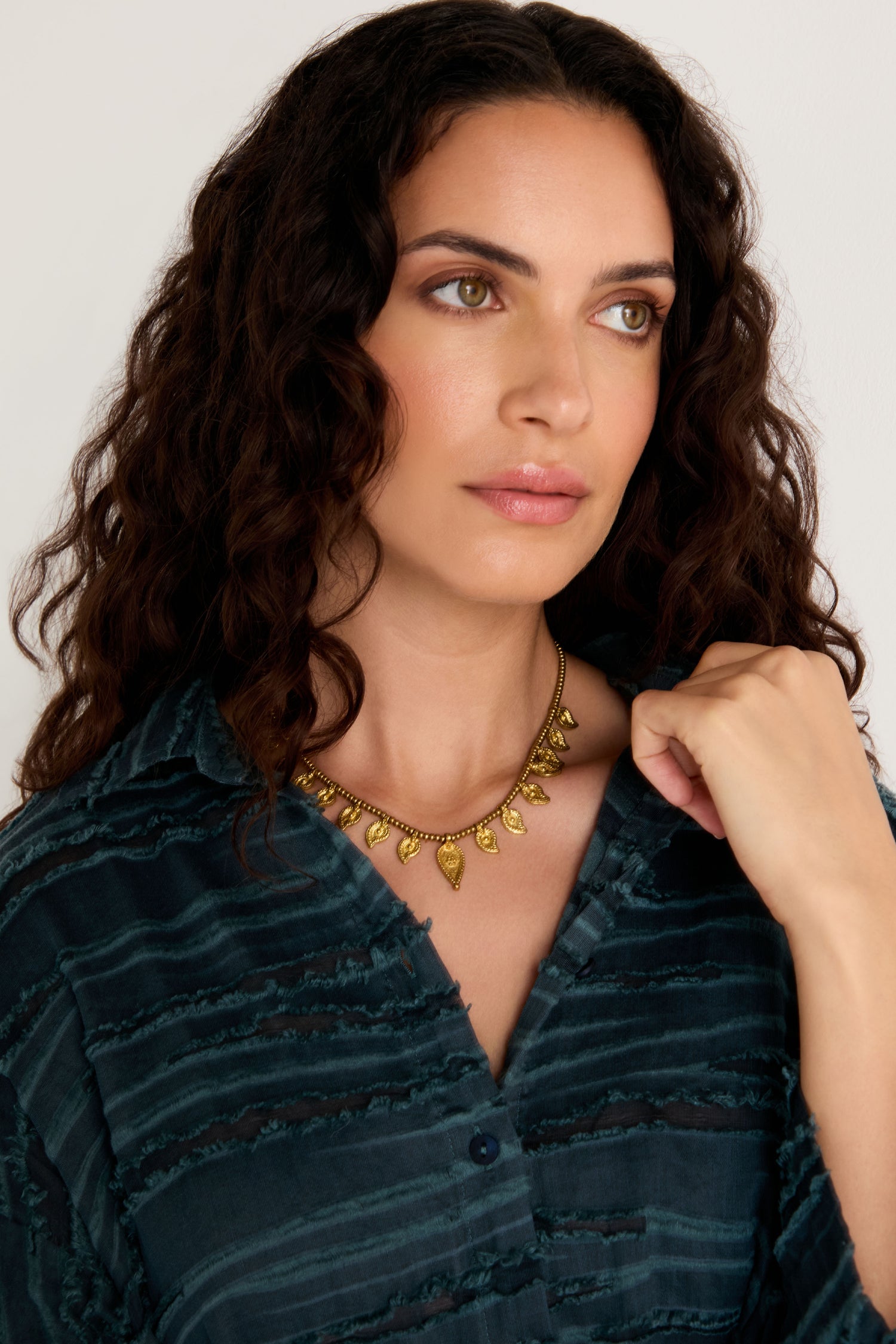 Woman with wavy brown hair, dressed in a dark textured blouse, wears the Paisley Necklace inspired by Indian metalwork. She gazes slightly to the side with a neutral expression.