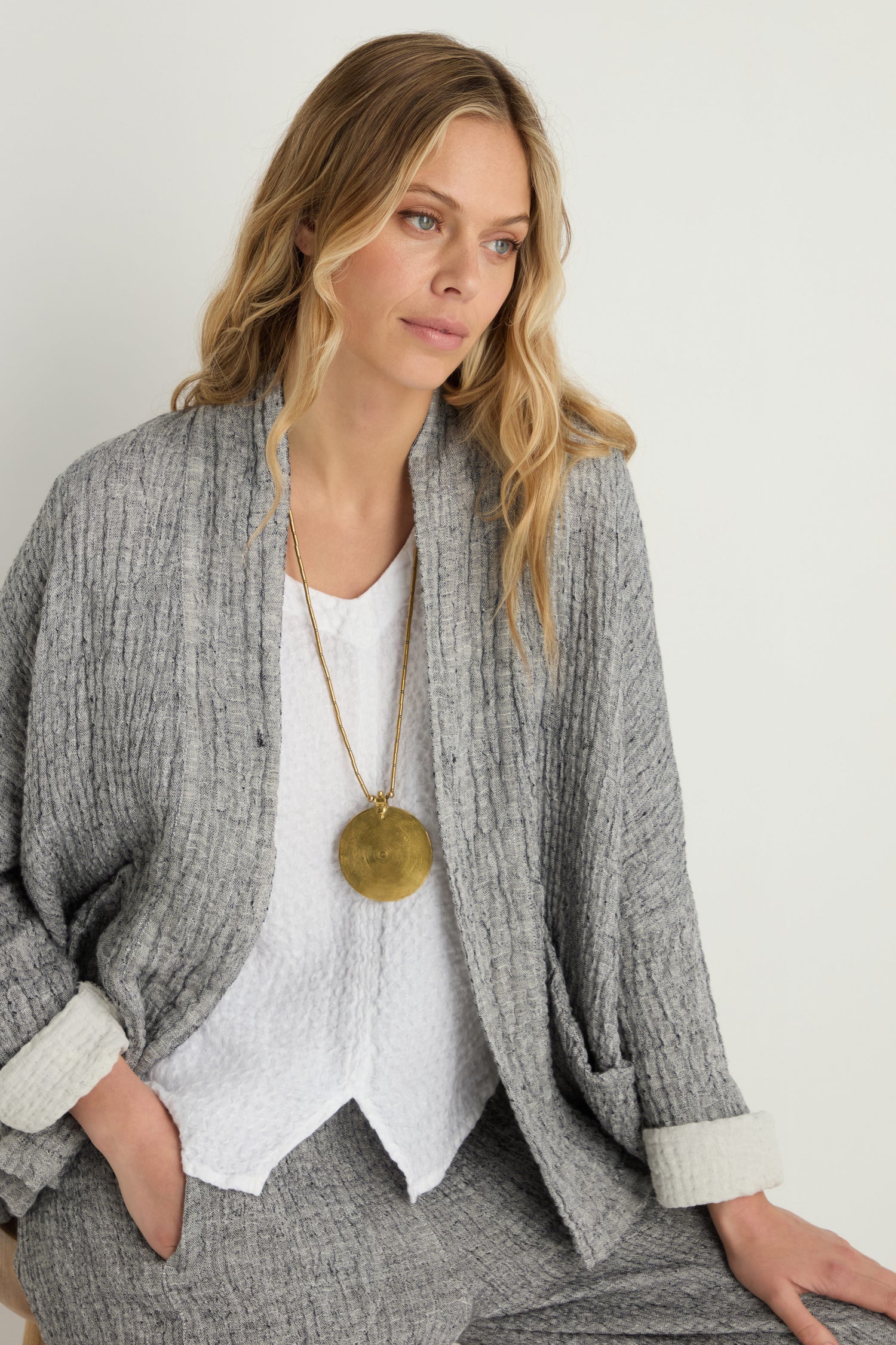 A woman with long blonde hair wears a textured gray cardigan and pants over a white top, accessorized with the Long Spiral Pendant statement necklace, seated against a plain light background.