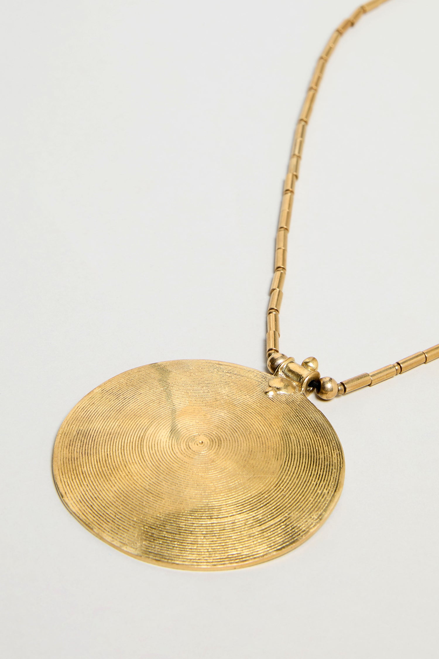 The Long Spiral Pendant is a gold necklace with a bold, flat spiral pendant adorned with fine concentric grooves, hanging from tubular chain links—a striking statement piece showcased on a plain background.