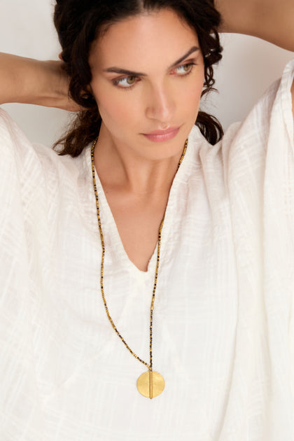 Woman in a white textured blouse models the handmade Long Mini Spiral Bead Pendant necklace, featuring black beads and a striking gold pendant, as she poses with arms raised behind her head and gazes to the side.