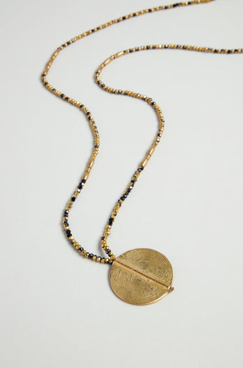 The Long Mini Spiral Bead Pendant, a beaded necklace in gold and black brass with a circular textured spiral bead, is displayed on a light surface.