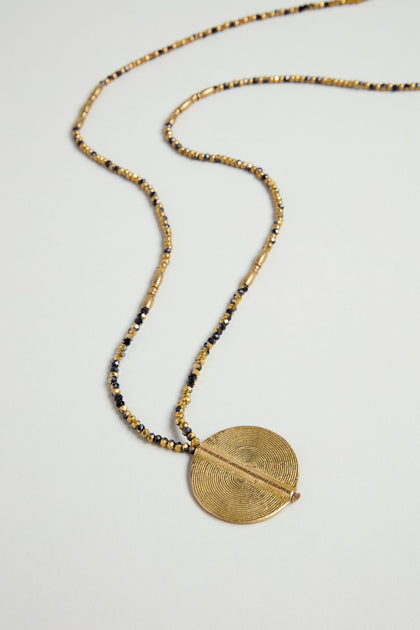 The Long Mini Spiral Bead Pendant, a beaded necklace in gold and black brass with a circular textured spiral bead, is displayed on a light surface.