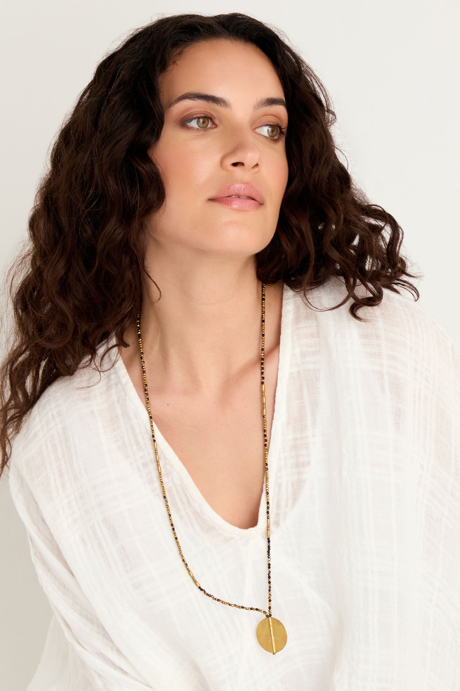 Woman with long curly hair wears a white textured top and the Long Mini Spiral Bead Pendant, a handmade Indian necklace with black bead detail, looking slightly to the side against a plain background.
