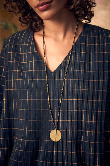 Long Mini Spiral Bead Pendant, a handmade Indian necklace with a brass bead spiral pendant worn over a dark pleated blouse with a gold grid pattern.