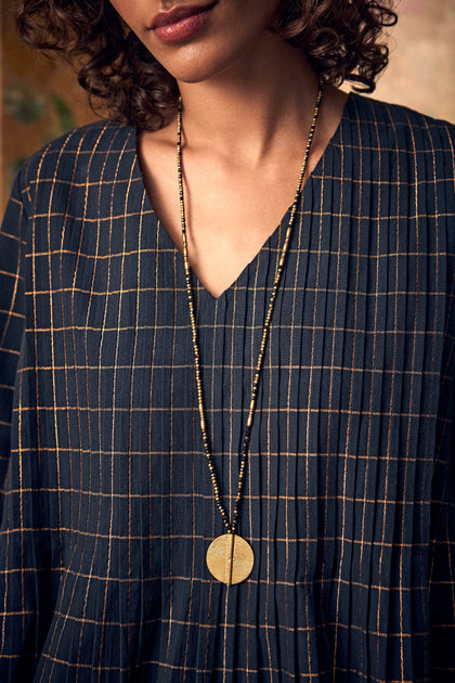 Long Mini Spiral Bead Pendant, a handmade Indian necklace with a brass bead spiral pendant worn over a dark pleated blouse with a gold grid pattern.