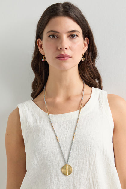 A woman with long dark hair, wearing a sleeveless white top, gold earrings, and an artisan-crafted necklace featuring the Moana Spiral Bead Pendant, stands against a plain background.