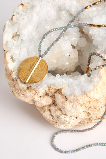 The Moana Spiral Bead Pendant, featuring elegant gold and small gray beads, is artfully draped over a large rough geode with a crystalline interior, highlighting exquisite artisan craftsmanship.