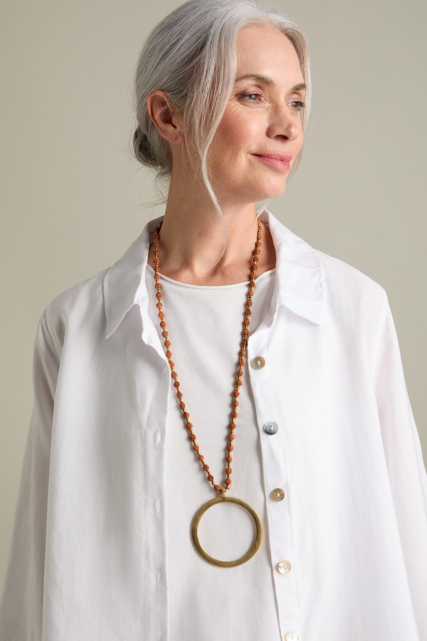 A woman with gray hair, wearing a white shirt and a long Circle Pendant Necklace, looks off to the side.