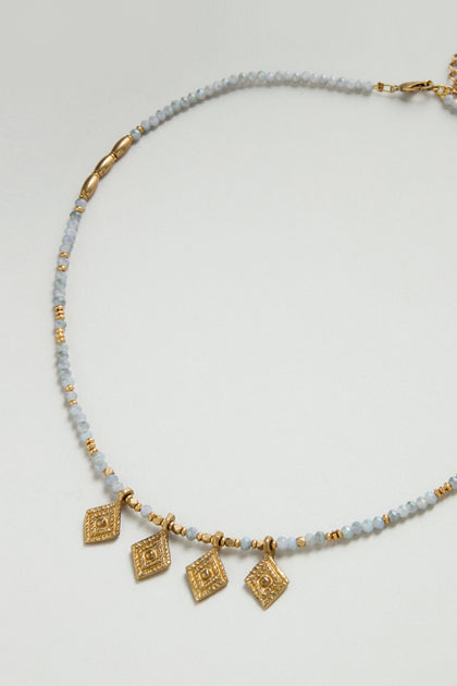 The Eanna Necklace features small light blue faceted glass beads, brass accents, and four evenly spaced gold diamond-shaped pendants at the front.