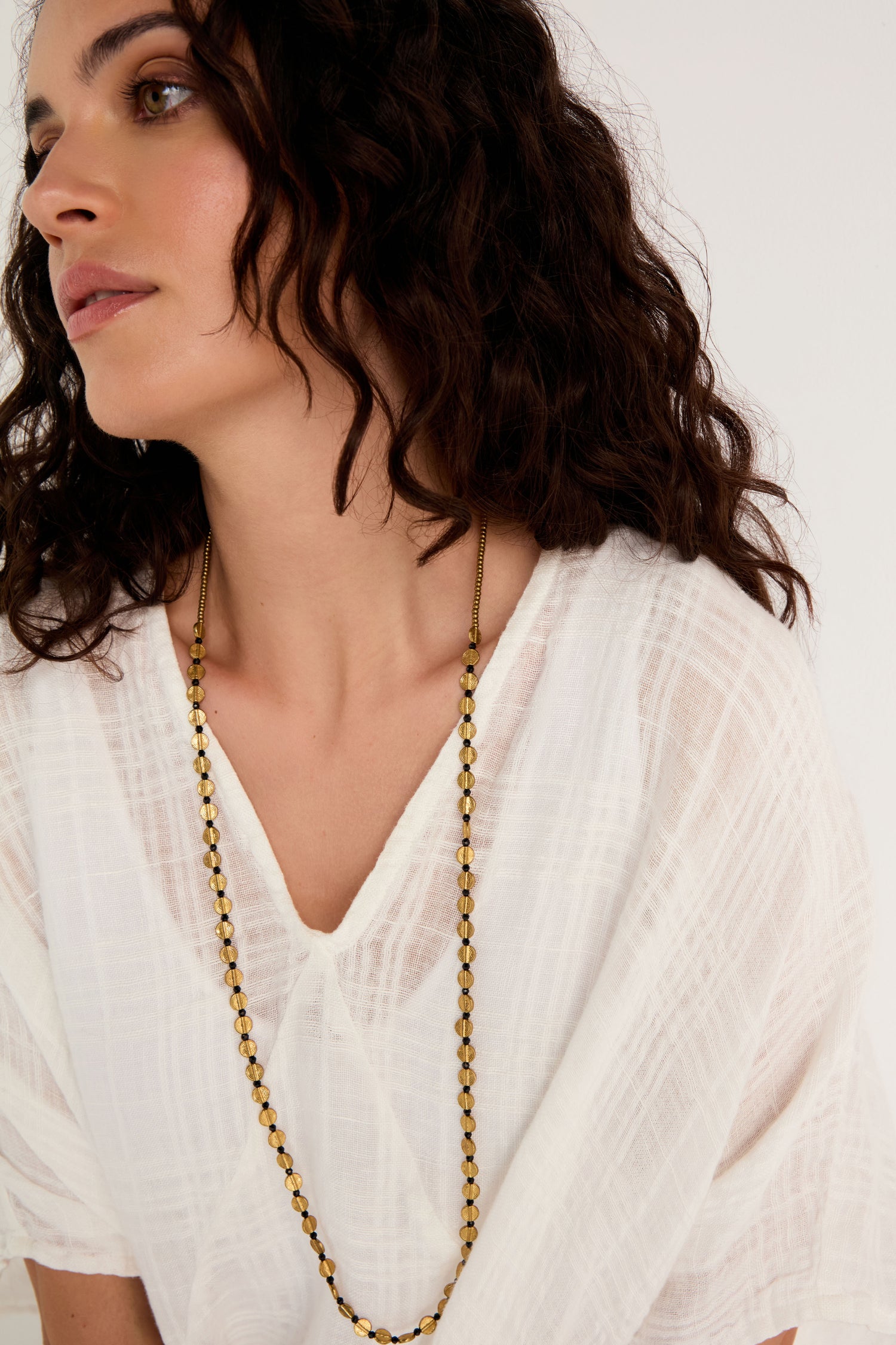 A woman with wavy dark hair wears a white textured blouse and the Mini Engraved Circles Bead Necklace, looking to the side against a plain background.