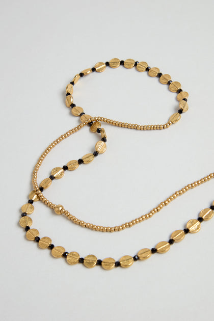 Two gold-toned beaded necklaces are displayed: one is a long brass bead strand, the other is the Mini Engraved Circles Bead Necklace featuring disc-shaped beads, black accents, and an engraved pendant.