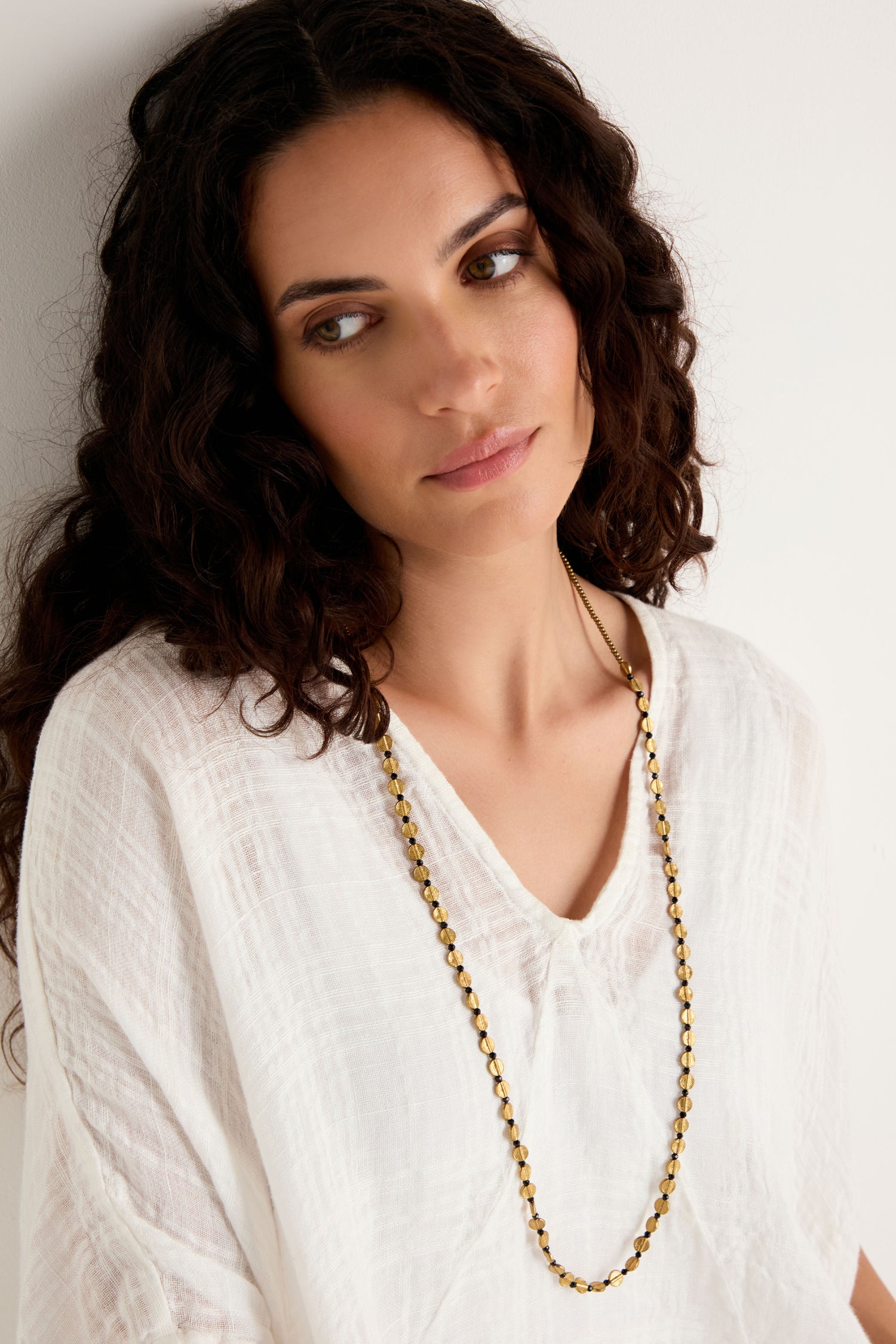 A woman with long curly hair wears a loose white textured top and the Mini Engraved Circles Bead Necklace, standing by a light-colored wall and gazing slightly to the side.