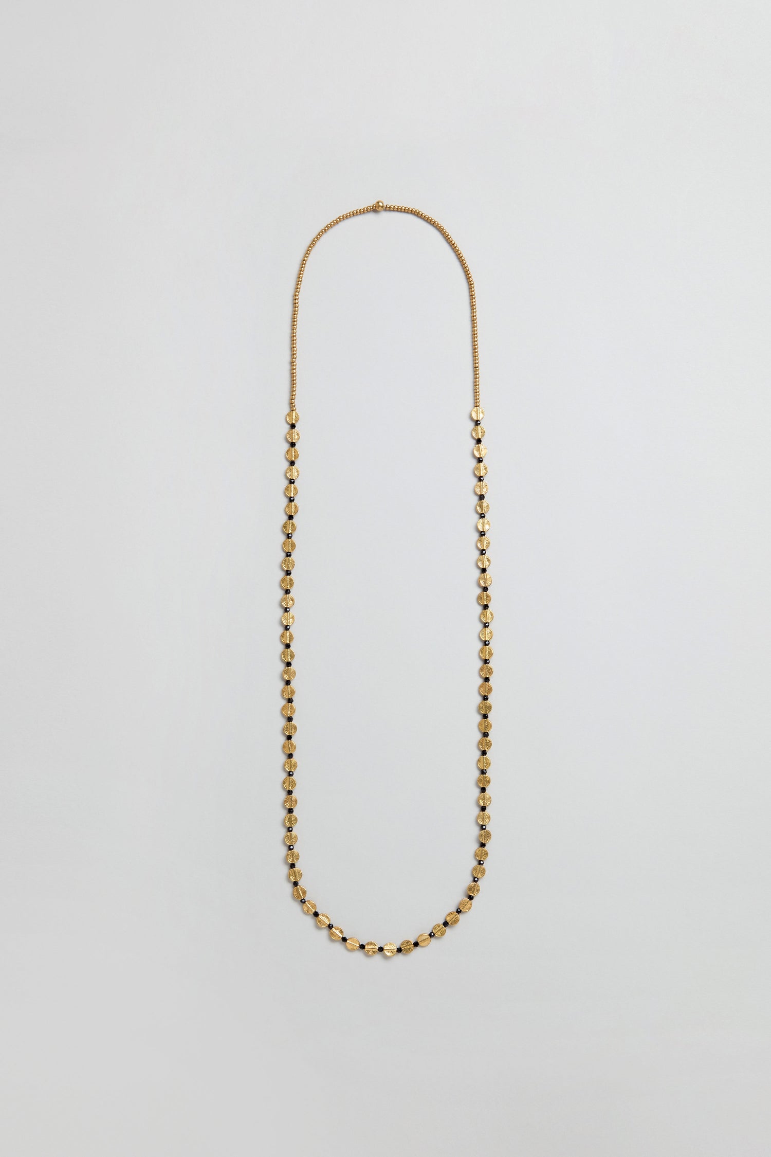 The Mini Engraved Circles Bead Necklace features small, evenly spaced gold oval beads and an elegant engraved disc pendant, displayed on a plain light gray background.