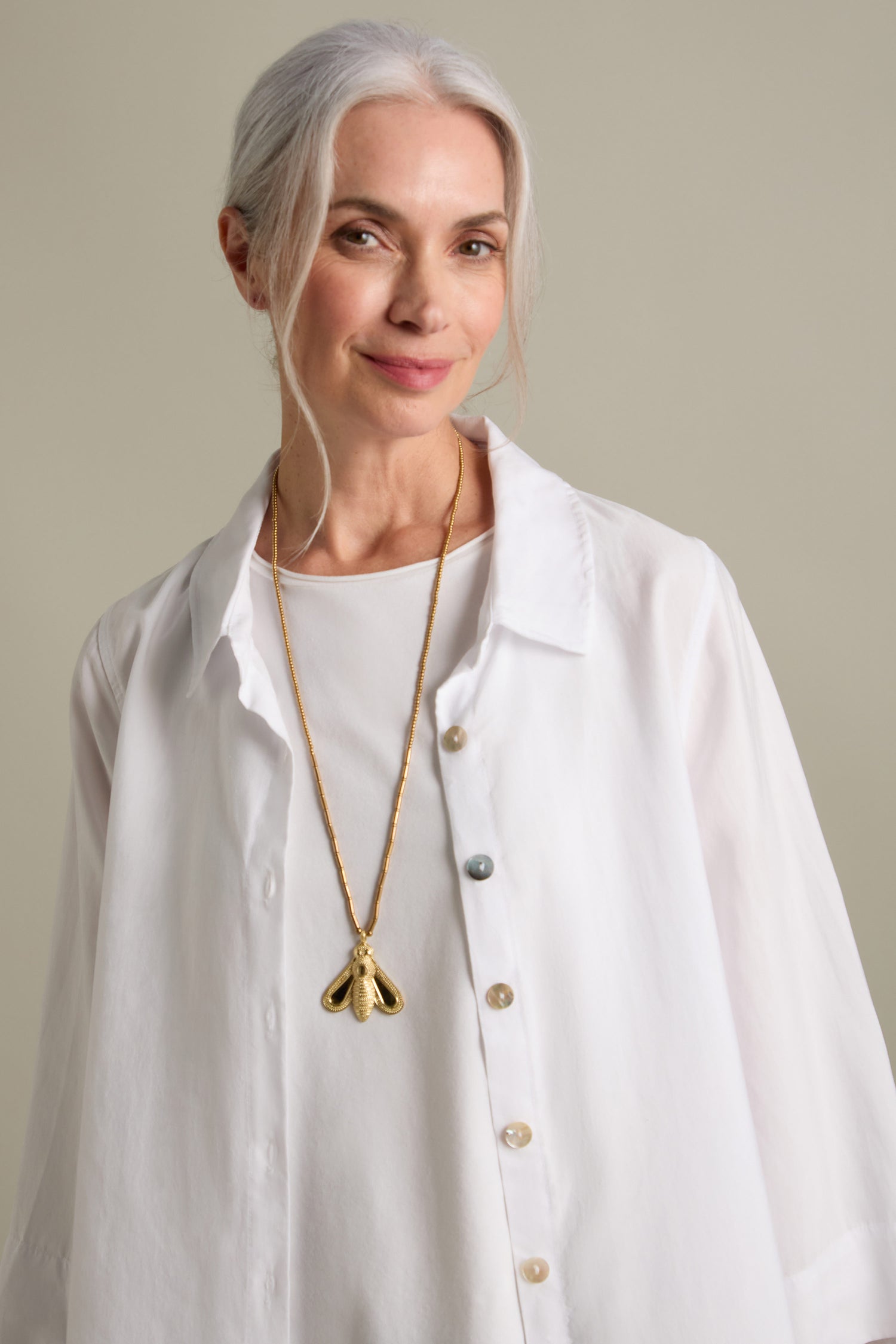 A woman with gray hair is smiling and wearing a white blouse. A long necklace adorned with gold beads and the Bumblebee Pendant rests gracefully on her chest. She has a relaxed expression and stands against a neutral background.