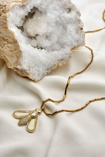 A Bumblebee Pendant necklace in gold lies on a white cloth, next to a large geode adorned with gleaming white crystals inside.