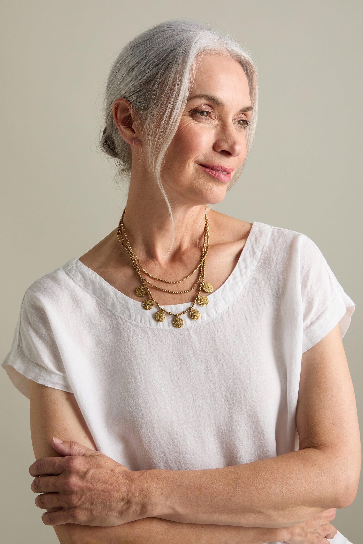 A woman with gray hair is wearing a white short-sleeved top and the Chakra Necklace, which features layered gold beads. She is standing with her arms crossed and looking slightly off to the side.