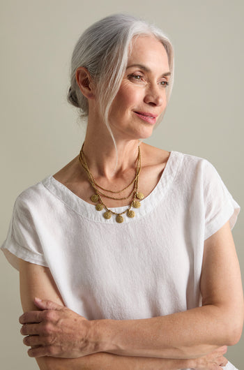 A woman with gray hair is wearing a white short-sleeved top and the Chakra Necklace, which features layered gold beads. She is standing with her arms crossed and looking slightly off to the side.