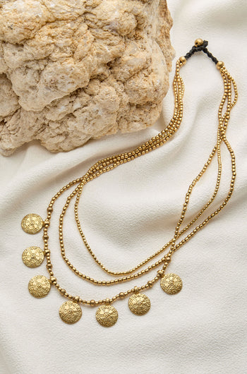 A Chakra Necklace featuring multiple layers of gold beads and round engraved chakra pendants, displayed on cream-colored fabric next to a textured rock.