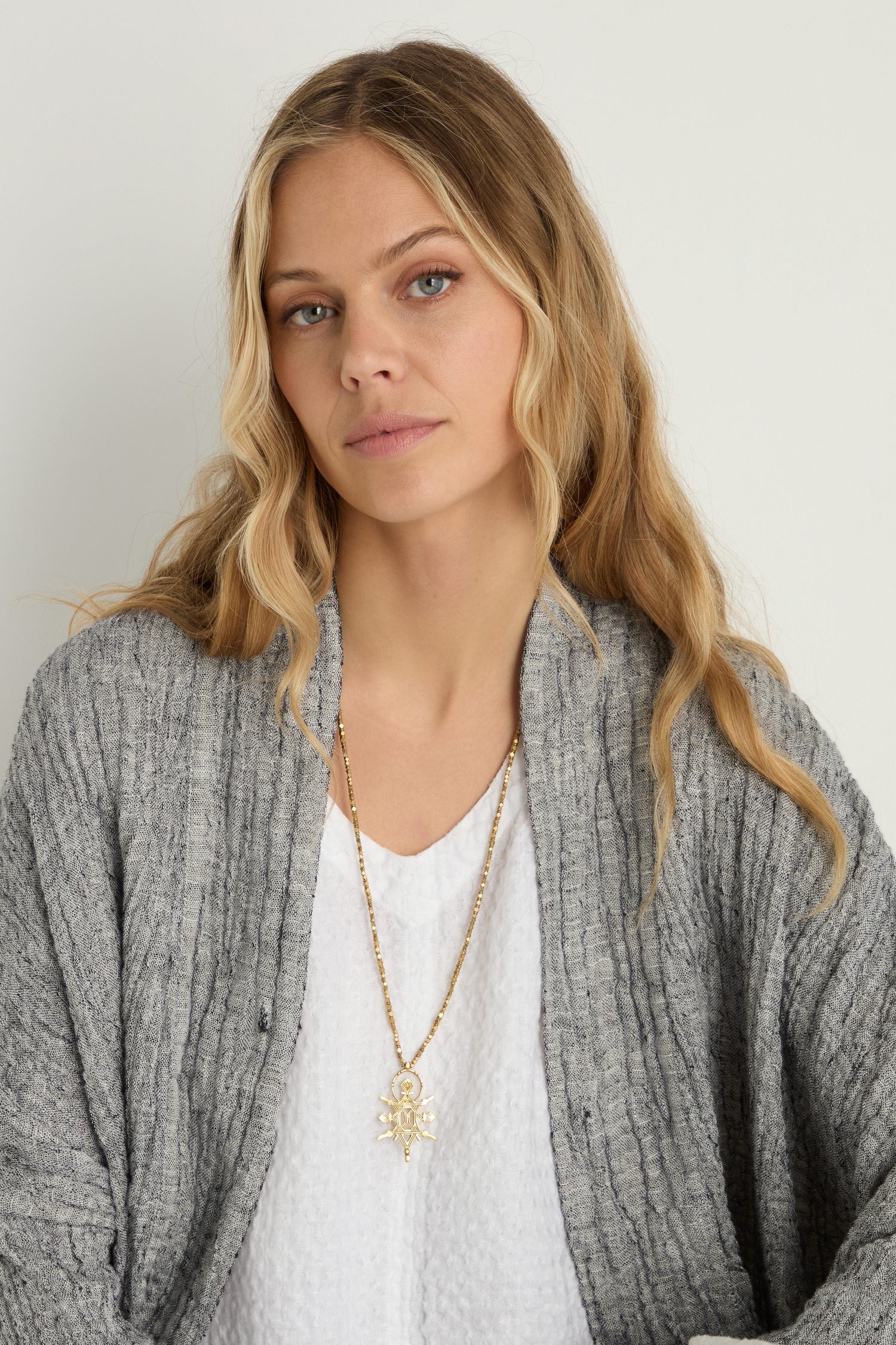 Woman with long blonde hair wears a grey cardigan, white textured top, and the Abalak Pendant—a geometric necklace on a gold-tone chain—while looking at the camera against a plain background.