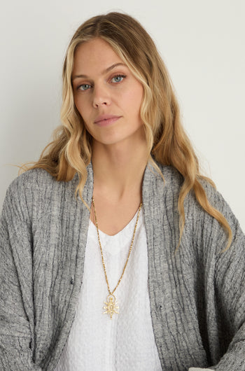Woman with long blonde hair wears a grey cardigan, white textured top, and the Abalak Pendant—a geometric necklace on a gold-tone chain—while looking at the camera against a plain background.