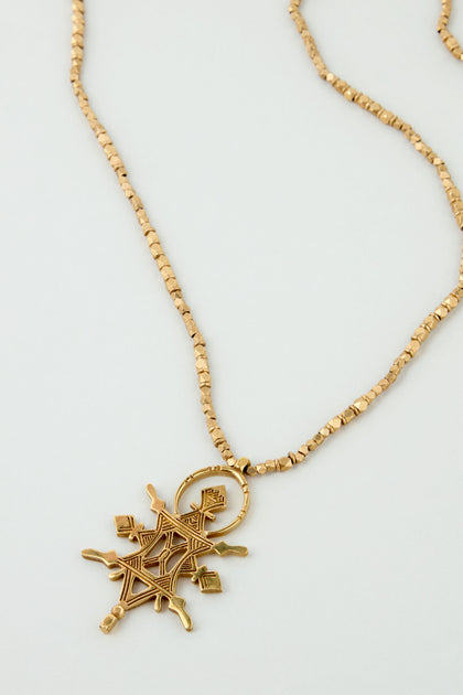 The Abalak Pendant is a gold tone chain necklace featuring a geometric, star-shaped pendant decorated with intricate patterns and small square accents.