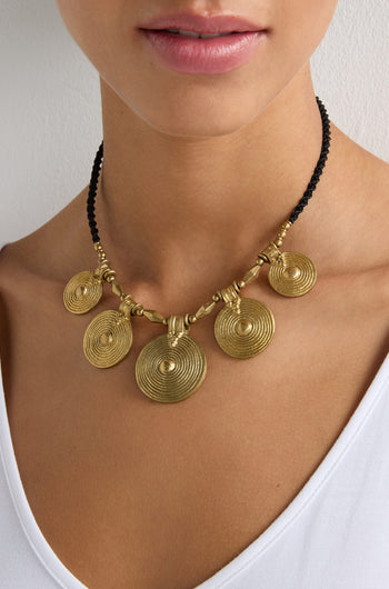 A person in a white top wears the Elissos Engraved Necklace, featuring five gold pendants on a black cord—highlighting contemporary elegance and exquisite handmade craftsmanship.