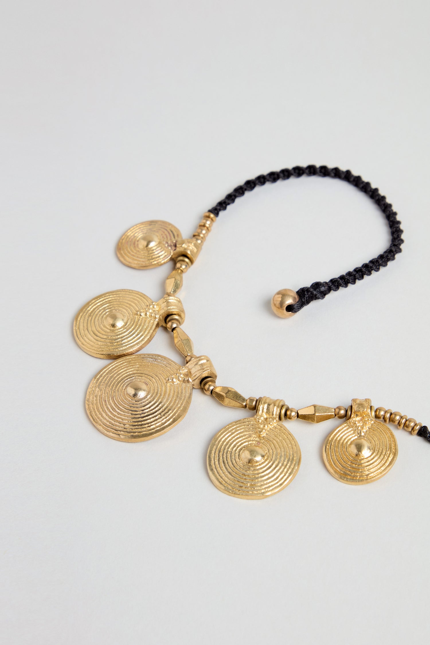 The Elissos Engraved Necklace features five round, gold-toned brass pendants with concentric circles, all handmade and attached to a black braided cord.