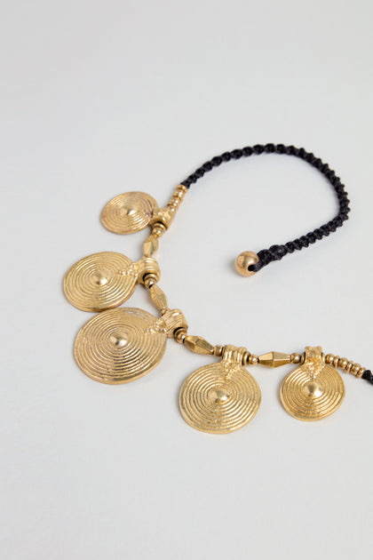 The Elissos Engraved Necklace features five round, gold-toned brass pendants with concentric circles, all handmade and attached to a black braided cord.