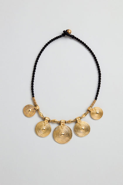 The Elissos Engraved Necklace features a black beaded cord and five evenly spaced brass pendants, each engraved with a circular spiral pattern—a striking handmade jewelry piece.