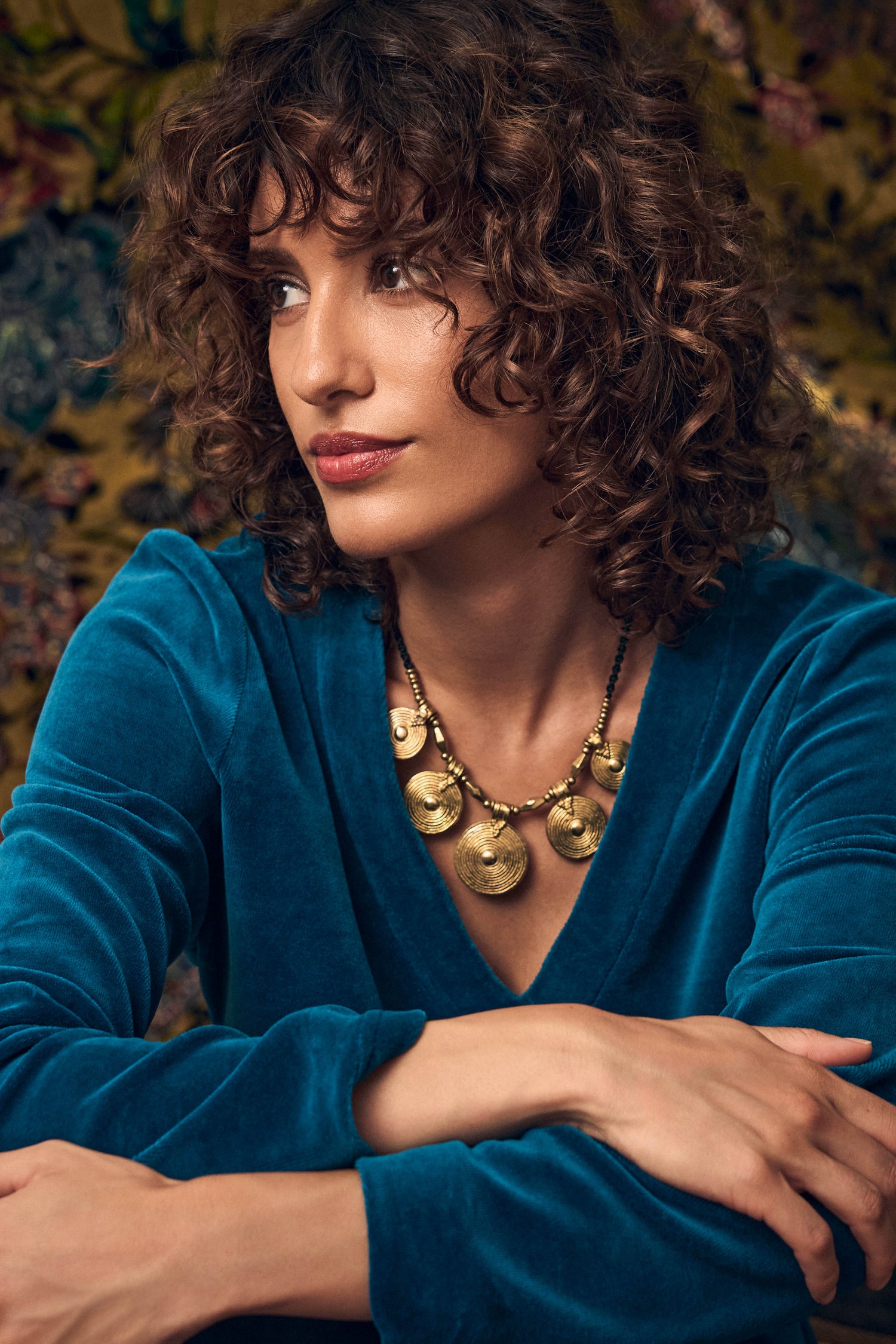 A person wearing an Elissos Engraved Necklace featuring gold-toned circular medallions on a dark beaded cord.