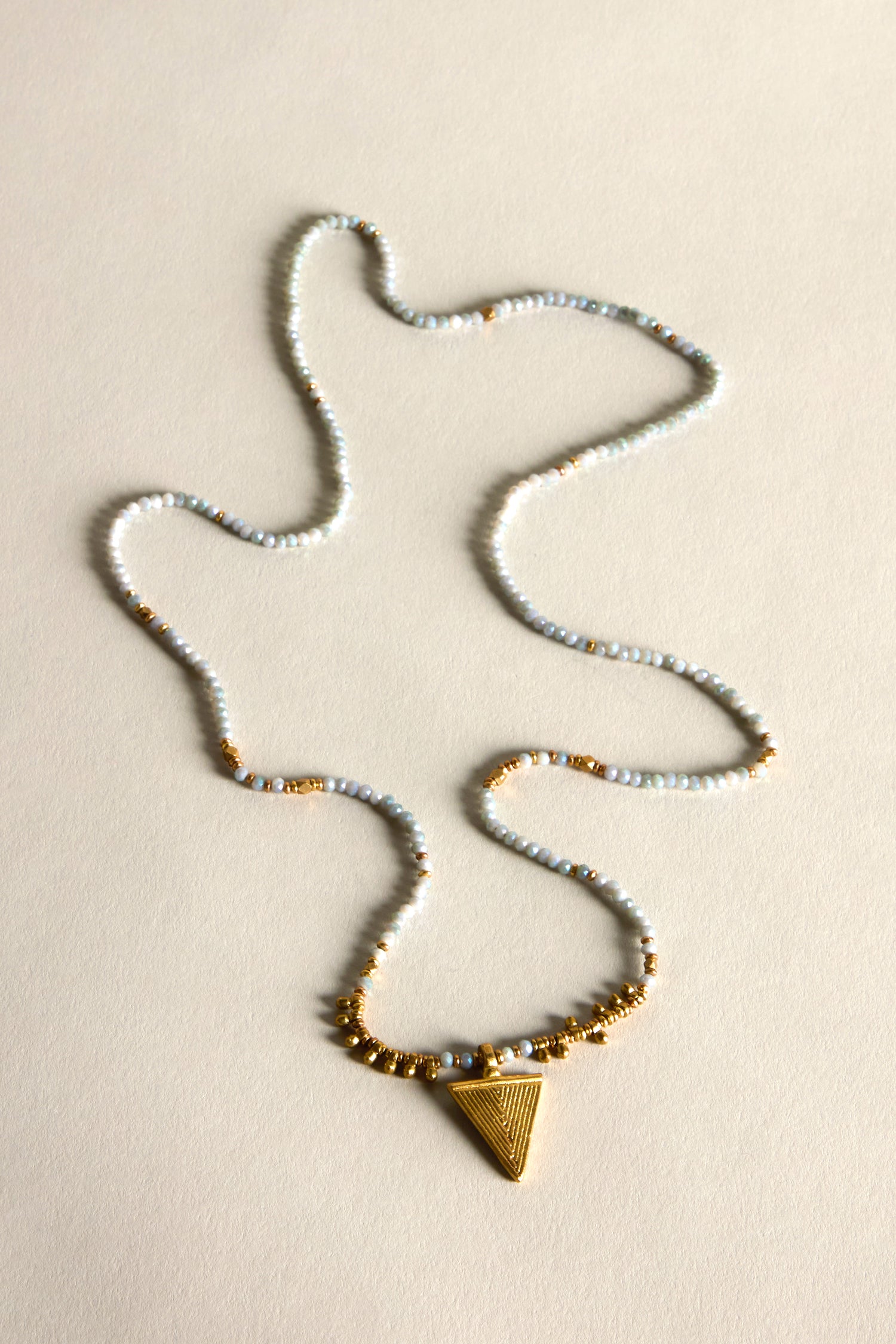 The Triskel Bead Pendant is a beaded necklace with blue and gold beads, a gold-tone arrowhead pendant, and pearlescent grey beads, set against a light background.