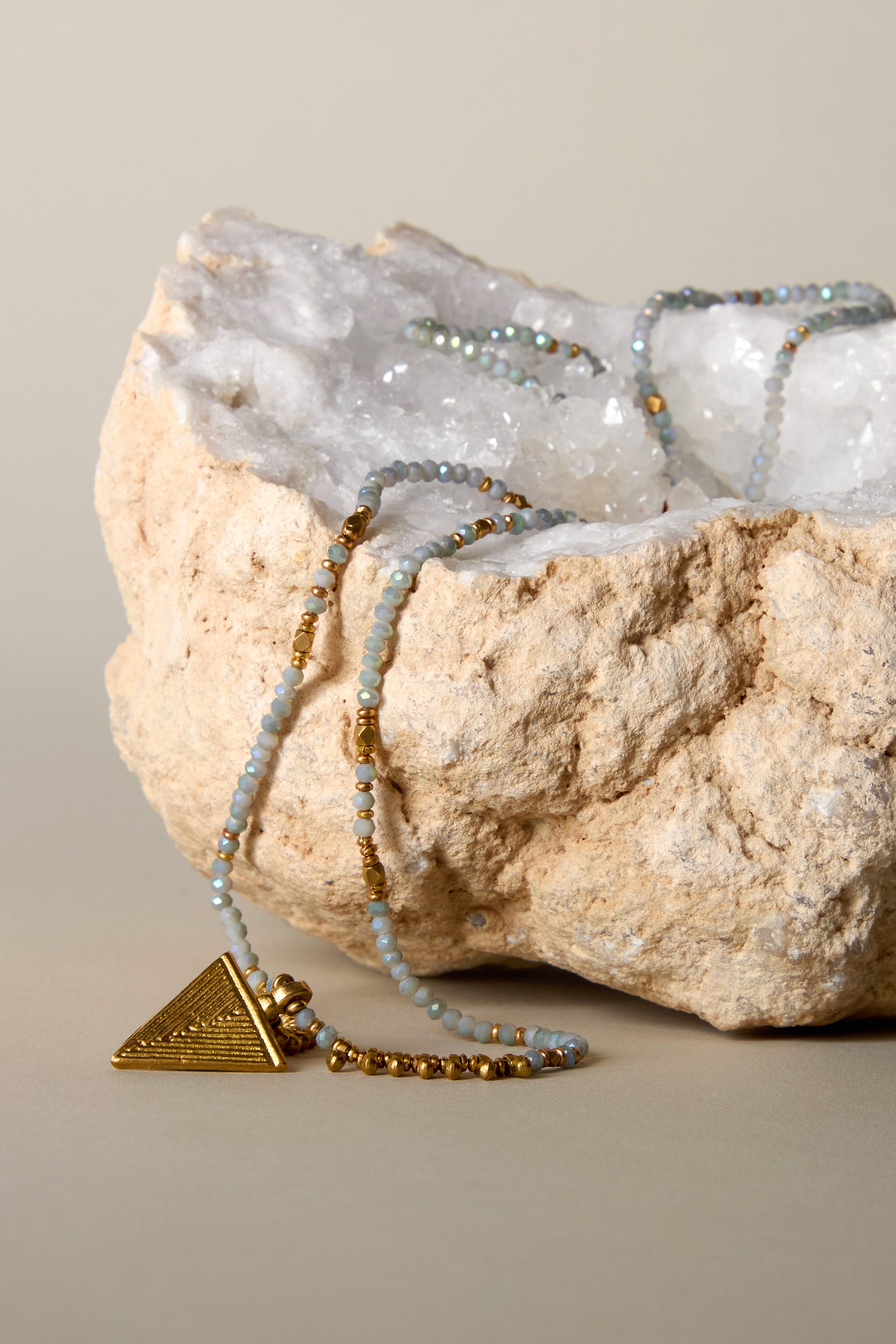 The Triskel Bead Pendant, featuring pearlescent grey beads and a gold-tone arrowhead pendant, elegantly drapes over a large textured rock with a white crystal interior.