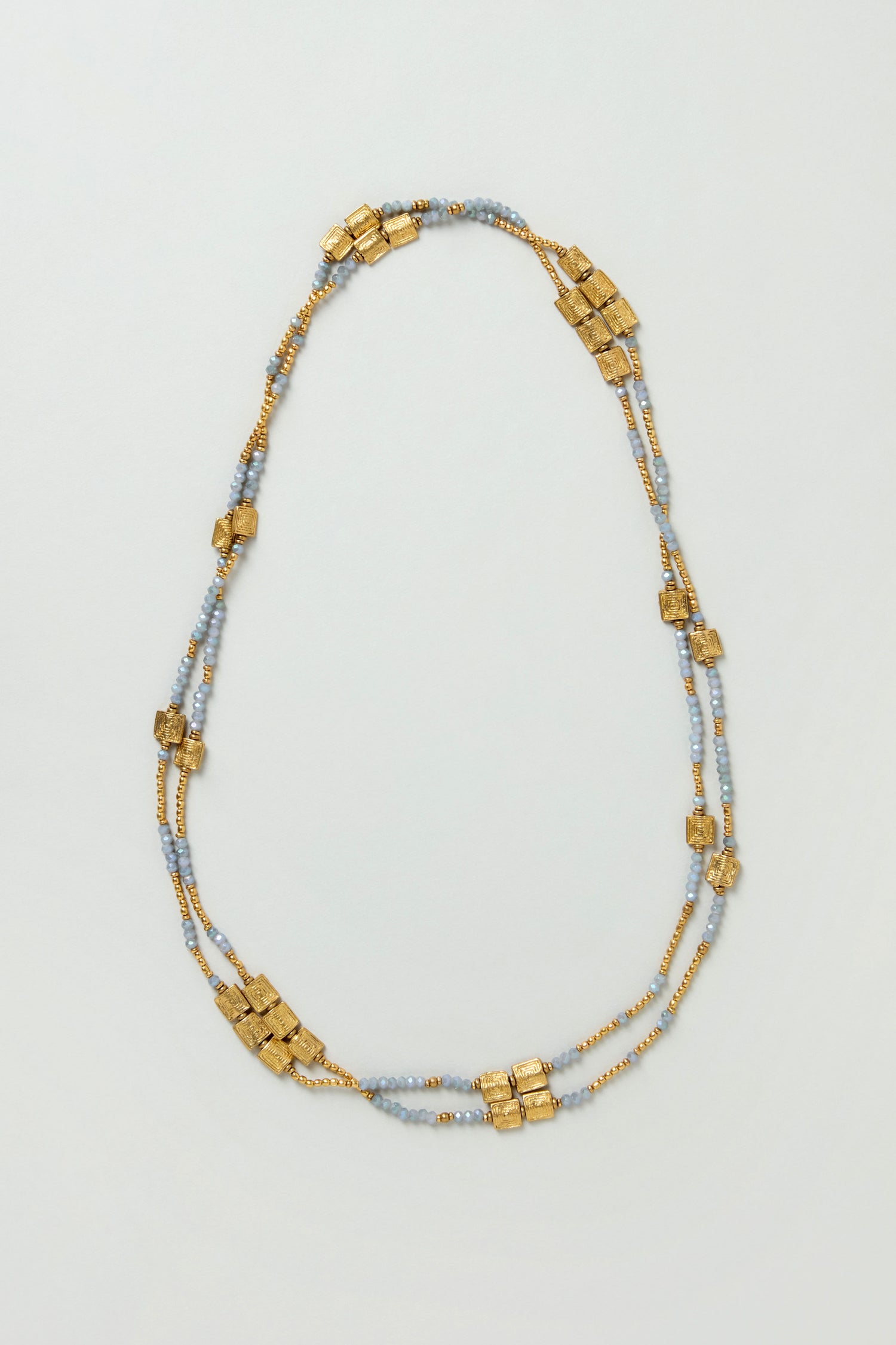 The Engraved Squares Bead Necklace is a layered design featuring small gold and light blue beads, plus clusters of larger brass and cut glass beads spaced along two strands for extra texture and shine.