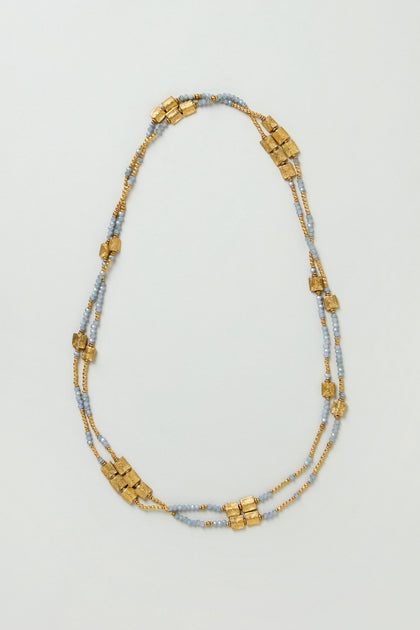 The Engraved Squares Bead Necklace is a layered design featuring small gold and light blue beads, plus clusters of larger brass and cut glass beads spaced along two strands for extra texture and shine.