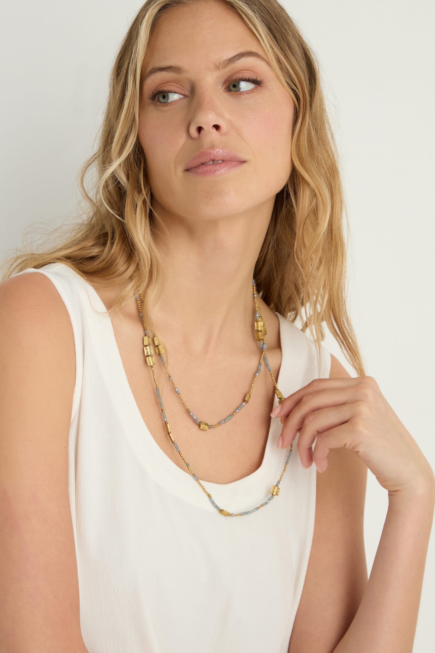 A woman with wavy blonde hair, in a sleeveless white top, showcases the Engraved Squares Bead Necklace made with brass and cut glass beads, gazing slightly to the side against a plain background.