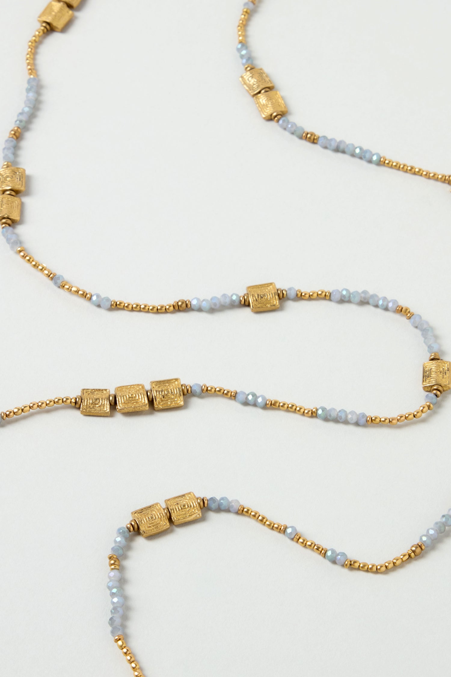 The Engraved Squares Bead Necklace is a long layered strand featuring small gold cylindrical beads, light blue round beads, and brass and cut glass accents arranged in a repeating pattern on a plain background.
