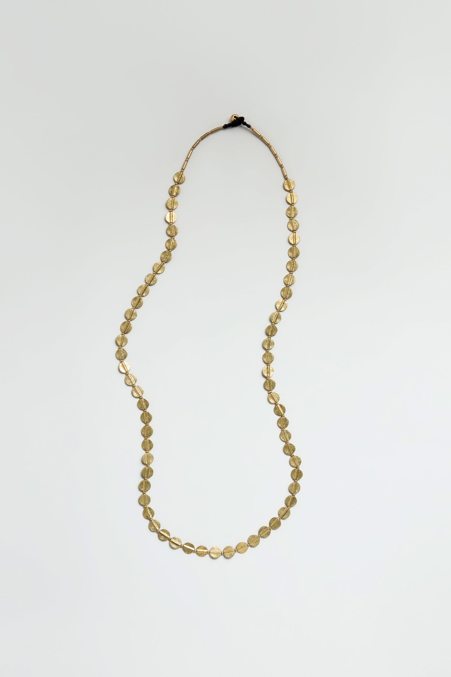 The Long Engraved Circles Necklace features polished brass, oval-shaped beads arranged evenly in a single strand, shown against a plain white background.
