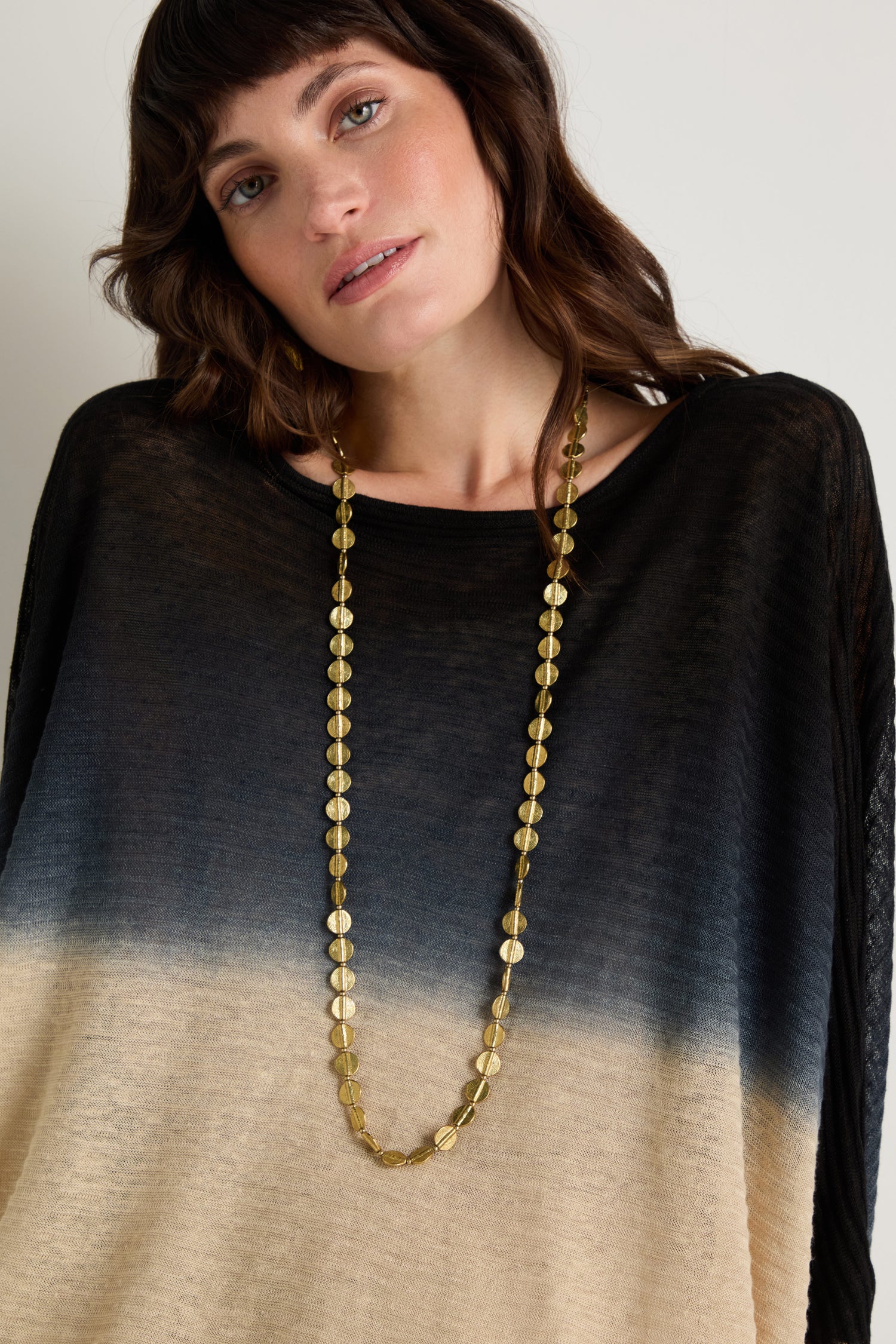 Woman with shoulder-length brown hair wearing a black and beige gradient sweater and the Long Engraved Circles Necklace, standing against a plain light background.