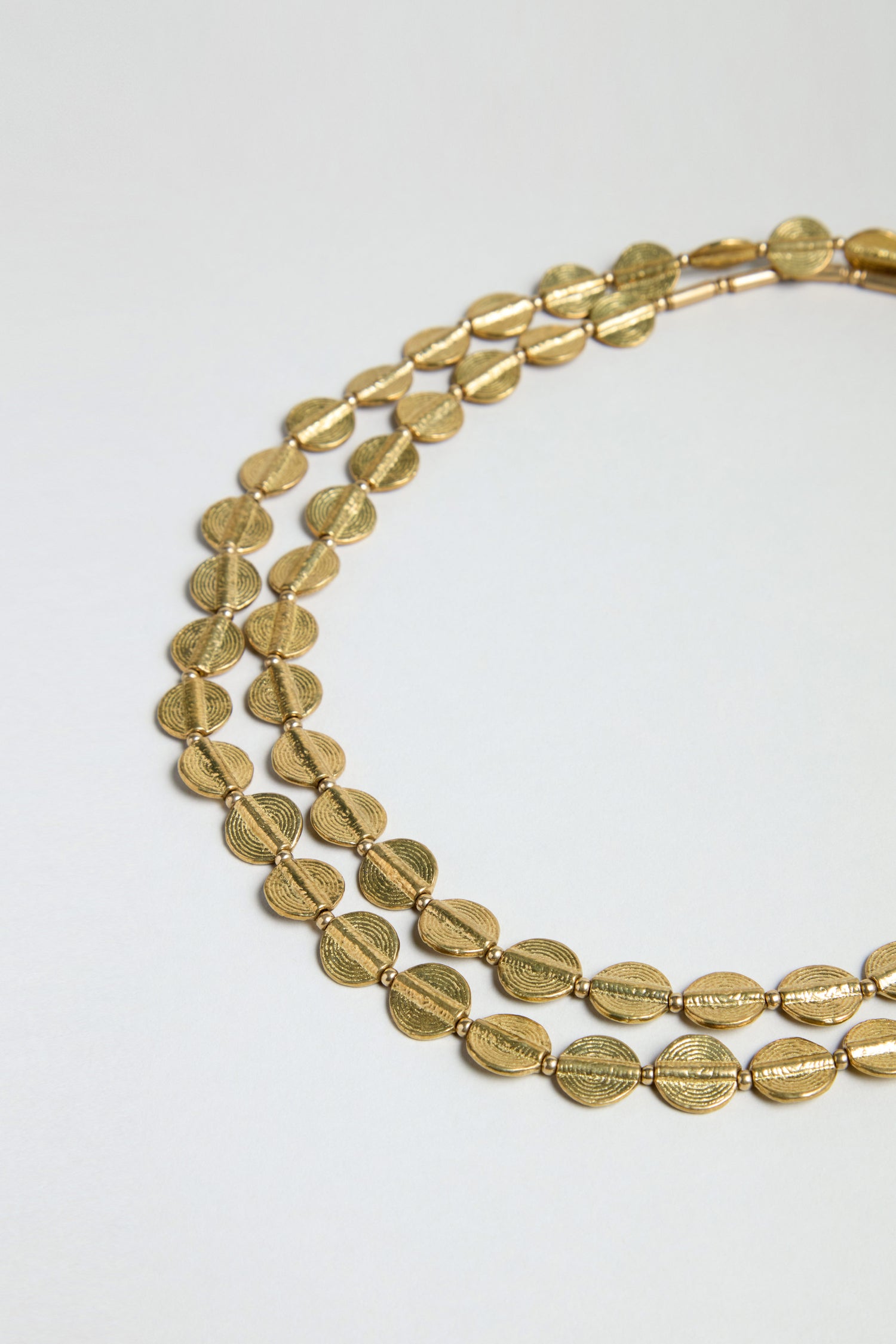 The Long Engraved Circles Necklace features two strands of gold-toned, oval beads with textured patterns, elegantly layered in a gentle curve on a simple, light background.