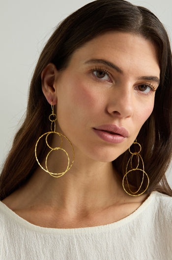 A woman with long brown hair wears Dharma Earrings—stylish gold-finished hoops from the Nataraj Collection—and a white top, gazing slightly to the side against a neutral background.