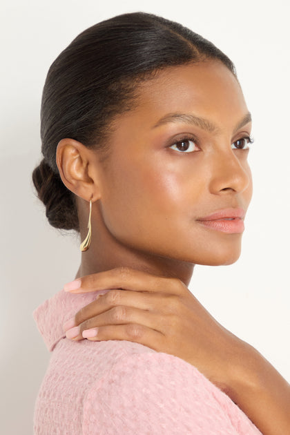 A woman with smooth skin and natural makeup wears Gilded Drop Earrings and a light pink textured top, posing with one hand resting on her shoulder against a plain background.
