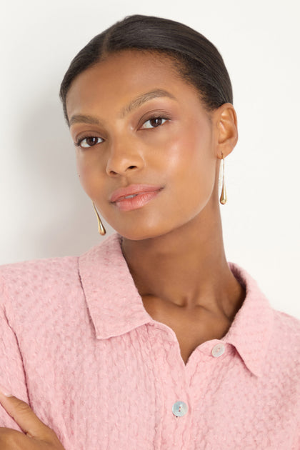A woman showcases the Gilded Drop Earrings while wearing a textured pink collared shirt, posed against a plain white background.