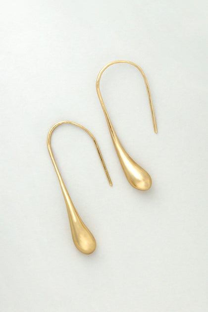 The Gilded Drop Earrings feature a teardrop shape and curved hooks, displayed against a light grey background.