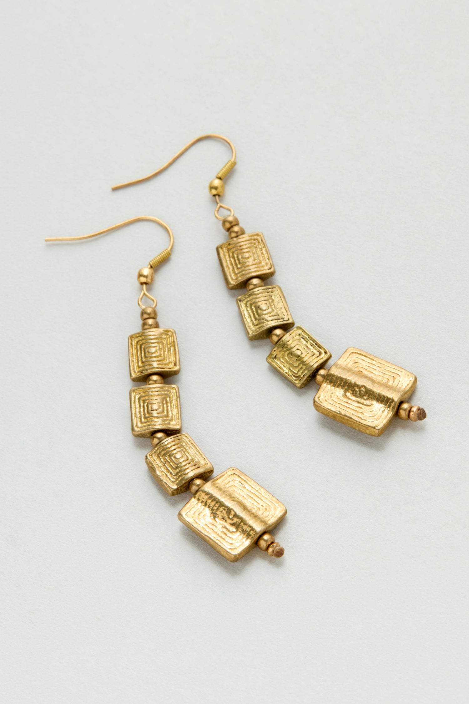 The Engraved Squares Earrings feature three stacked, textured square beads in gold-tone brass with hook ear wires, arranged vertically and showcased against a plain background.