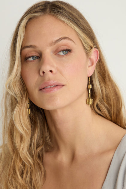 A woman with long, wavy blonde hair wears Engraved Squares Earrings—sculptural gold geometric jewelry—and a light grey top, looking slightly to the side against a neutral background.