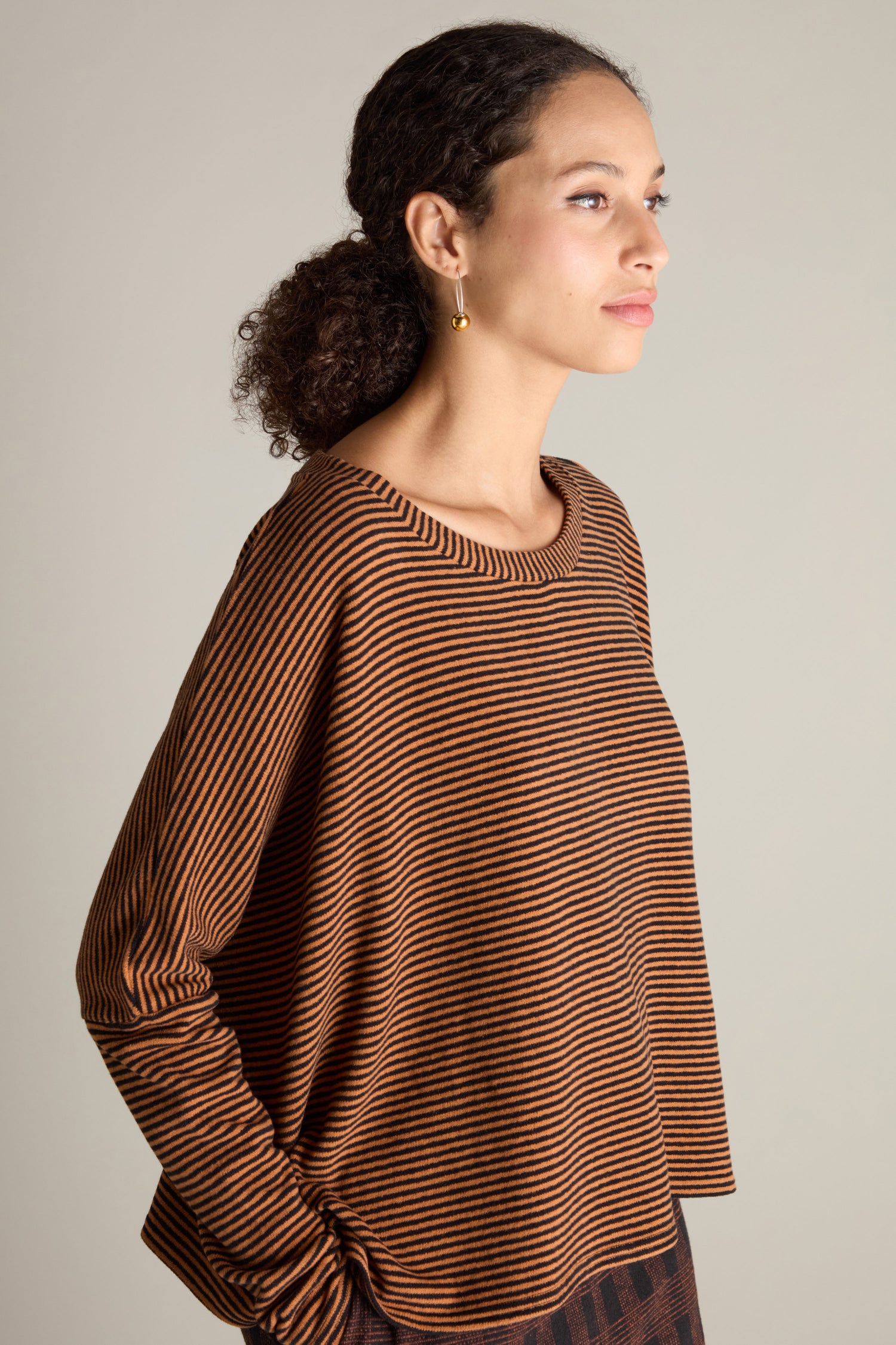 A person is photographed in profile, wearing a Neirami Mini Stripe Boxy Top in brown and black. They have curly hair pulled back and are accessorized with a gold earring, showcasing this versatile wardrobe essential in style.