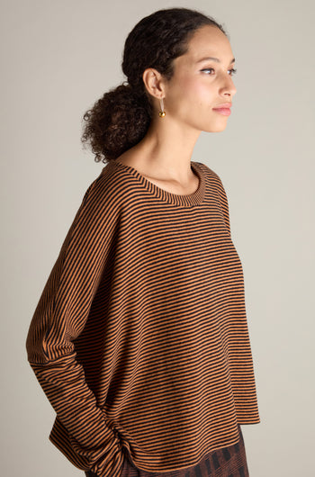 A person is photographed in profile, wearing a Neirami Mini Stripe Boxy Top in brown and black. They have curly hair pulled back and are accessorized with a gold earring, showcasing this versatile wardrobe essential in style.