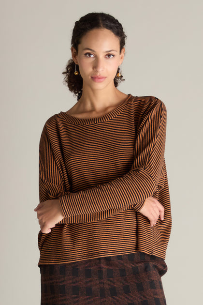 A person with curly hair wearing a Mini Stripe Boxy Top and a checkered skirt stands with folded arms against a plain background, showcasing a versatile wardrobe essential.